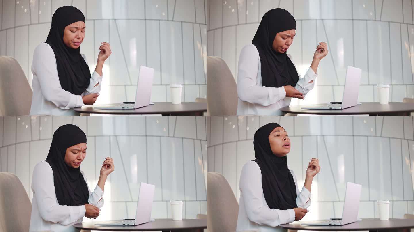 戴着黑色头巾的多种族妇女由于在现代办公室久坐不动而肘部疼痛。悲伤的女经理坐在办公桌前，带着无线笔记本