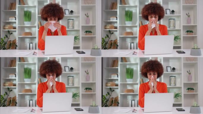 悲伤的非裔美国女人坐在办公桌前，拿着现代笔记本电脑，用餐巾打喷嚏。穿着橙色罩衫的年轻女办公室职员在季