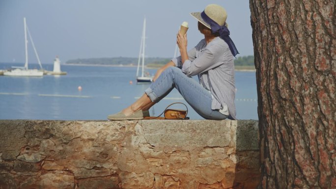 克罗地亚港口的挡土墙上，一名女游客一边吃冰淇淋一边放松