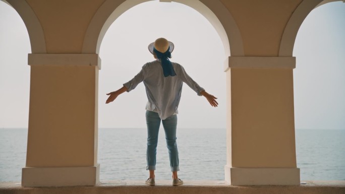 在克罗地亚晴朗的天空下，女游客站在海边柱廊的挡土墙上，伸开双臂