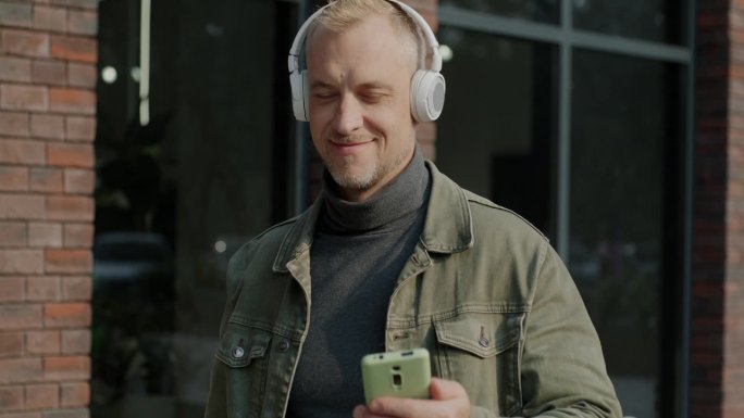 一个快乐的家伙戴着无线耳机站在城市街道上听音乐的慢动作