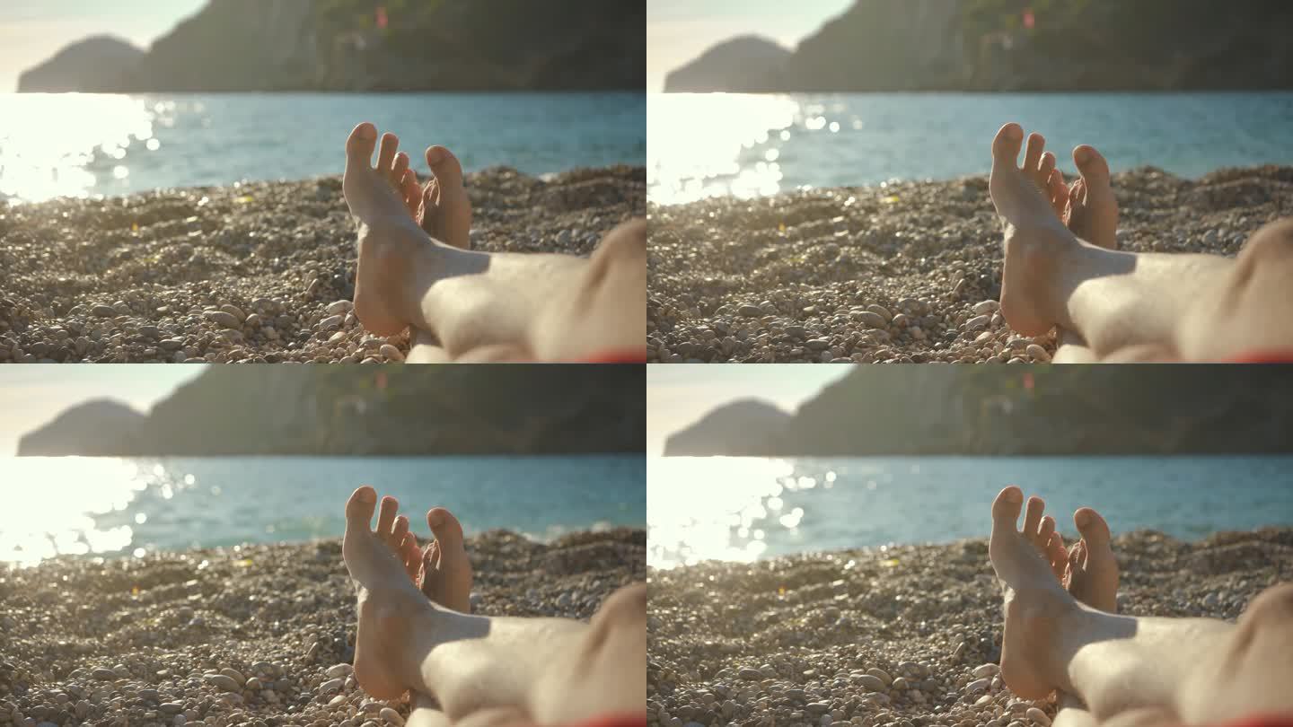 年轻人躺在卵石滩上休息，只看得见交叉的光脚，迷离的午后阳光照亮了平静的海面，背景是岩石嶙峋的悬崖