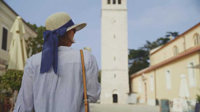 阳光明媚的一天，克罗地亚罗维尼，一名兴高采烈的女游客一边朝大教堂塔楼走去，一边吃着冰淇淋