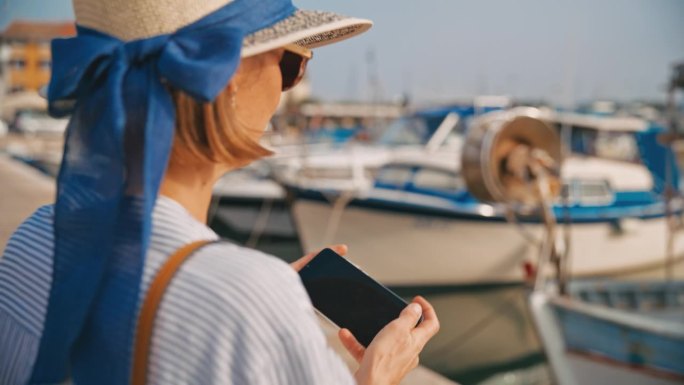一名女游客坐在克罗地亚村庄的码头柱上，用智能手机拍摄港口
