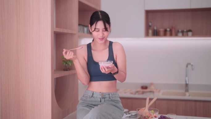 穿着运动服的亚洲女运动员在舒适的厨房里品尝着清爽的酸奶。她崇尚健康的生活方式，用有益健康的食物滋养自