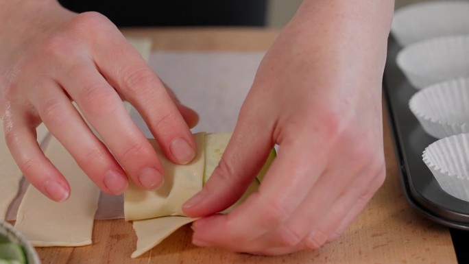 在木桌上，一个女孩用手把西葫芦和奶酪磨碎的酥皮卷成玫瑰花形，然后放在松饼烤盘里。正面，特写