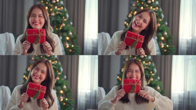 快乐迷人的亚洲女人拿着一份红色的礼物对着镜头。微笑的女人坐在一棵装饰着圣诞装饰品的圣诞树旁，平安夜，