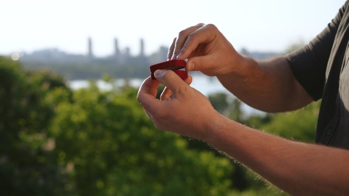 一个男人合上装有戒指的盒子，捏在手里，背景是河边的一座城市。准备求婚。