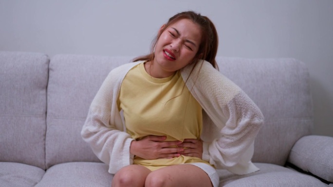 患病的亚洲妇女因经期(经前症候群)而腹部剧烈疼痛。坐在沙发上，食物中毒引起胃痛，腹痛，消化问题，胃炎