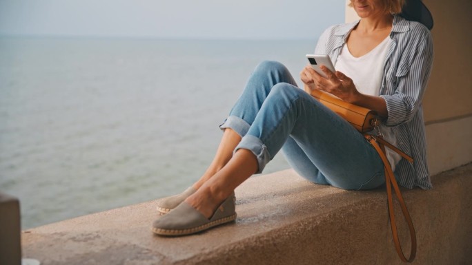 克罗地亚，一名女游客坐在海边的挡土墙上使用智能手机