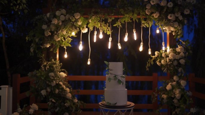 户外聚会上的白色大蛋糕，上面有闪亮的花环和装饰花拱。
