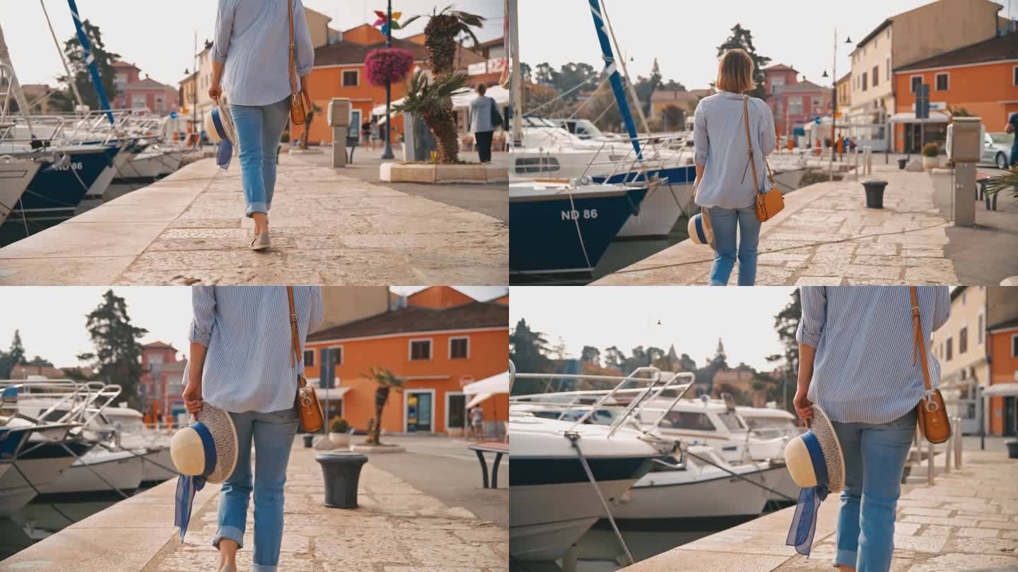女游客带着吊带包和遮阳帽走在克罗地亚村庄的码头上，探索港口的后视图