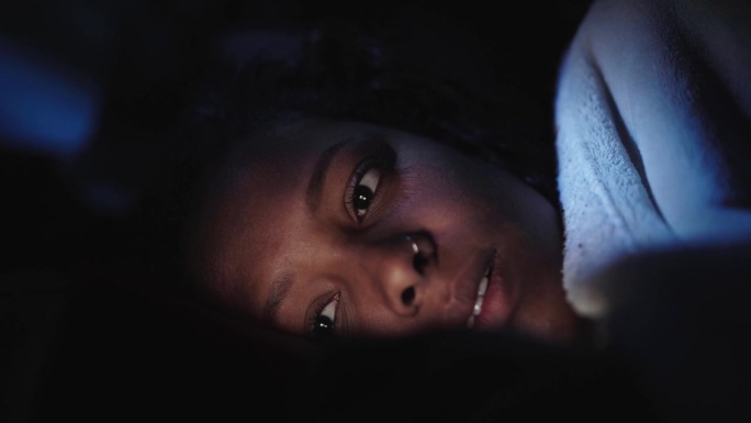 年轻的非裔美国妇女在黑暗中放松地看手机的特写。脸照亮。