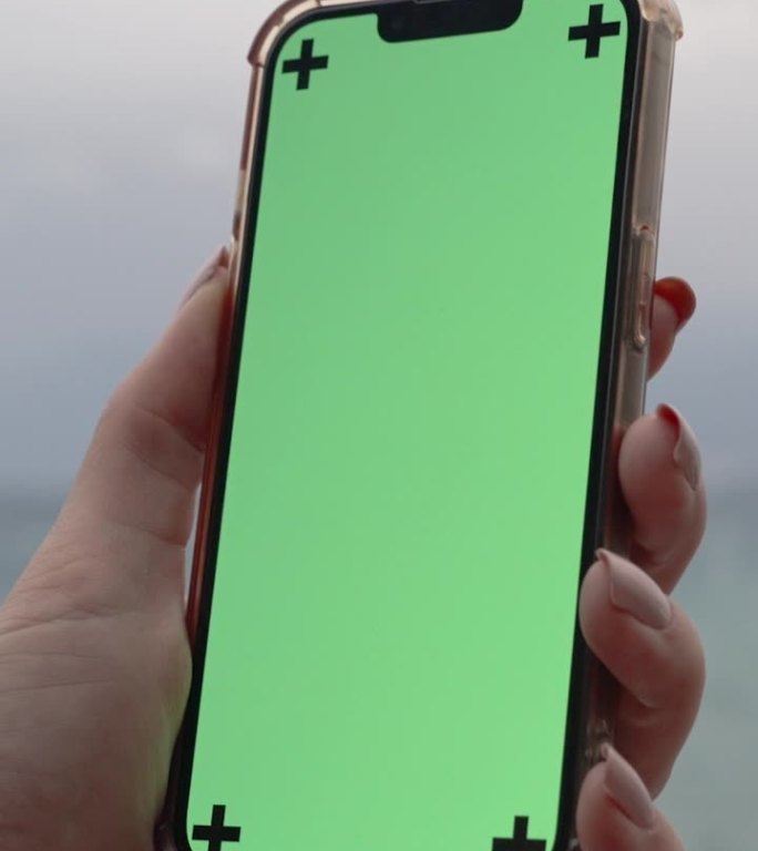 垂直视频。一位女性手持一部手机，上面有一个空白的绿色屏幕和标记，背景是大海和阴天。