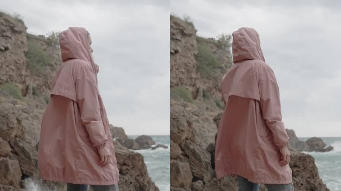 垂直视频。一个穿着粉色雨衣的女人站在尖锐的岩石上，看着大浪冲击着她身边的石头。海上风暴。海上风暴。