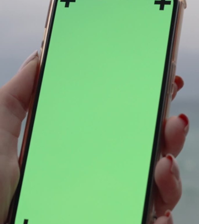 垂直视频。特写，放大的女性手拿着手机与绿色屏幕的模拟。在背景中，暴风雨的大海和山脉。
