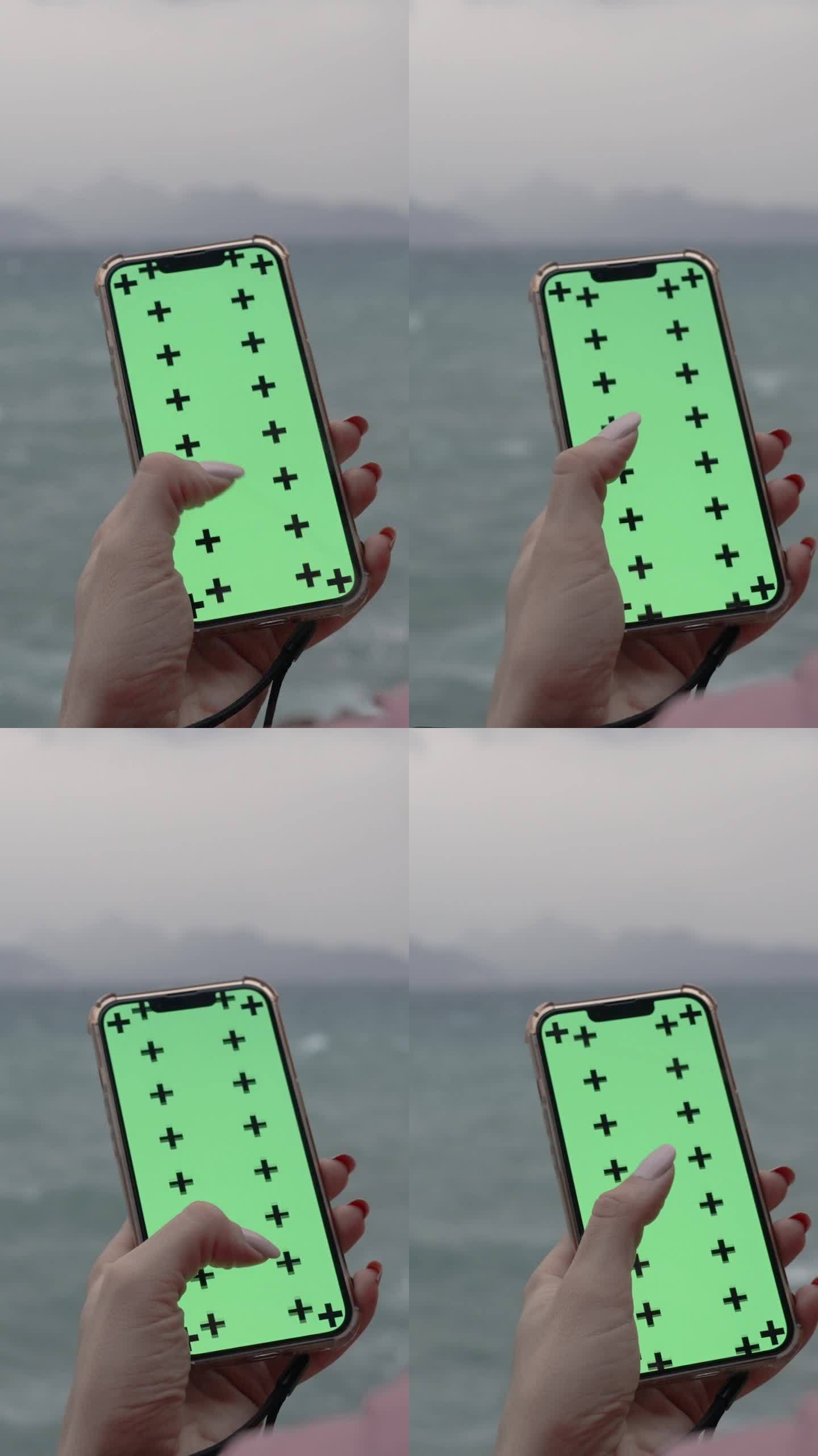 垂直视频。从一个女人的头后面看，她在智能手机上滚动着一些东西，绿色的屏幕是一个模型，背景是海边阴天的