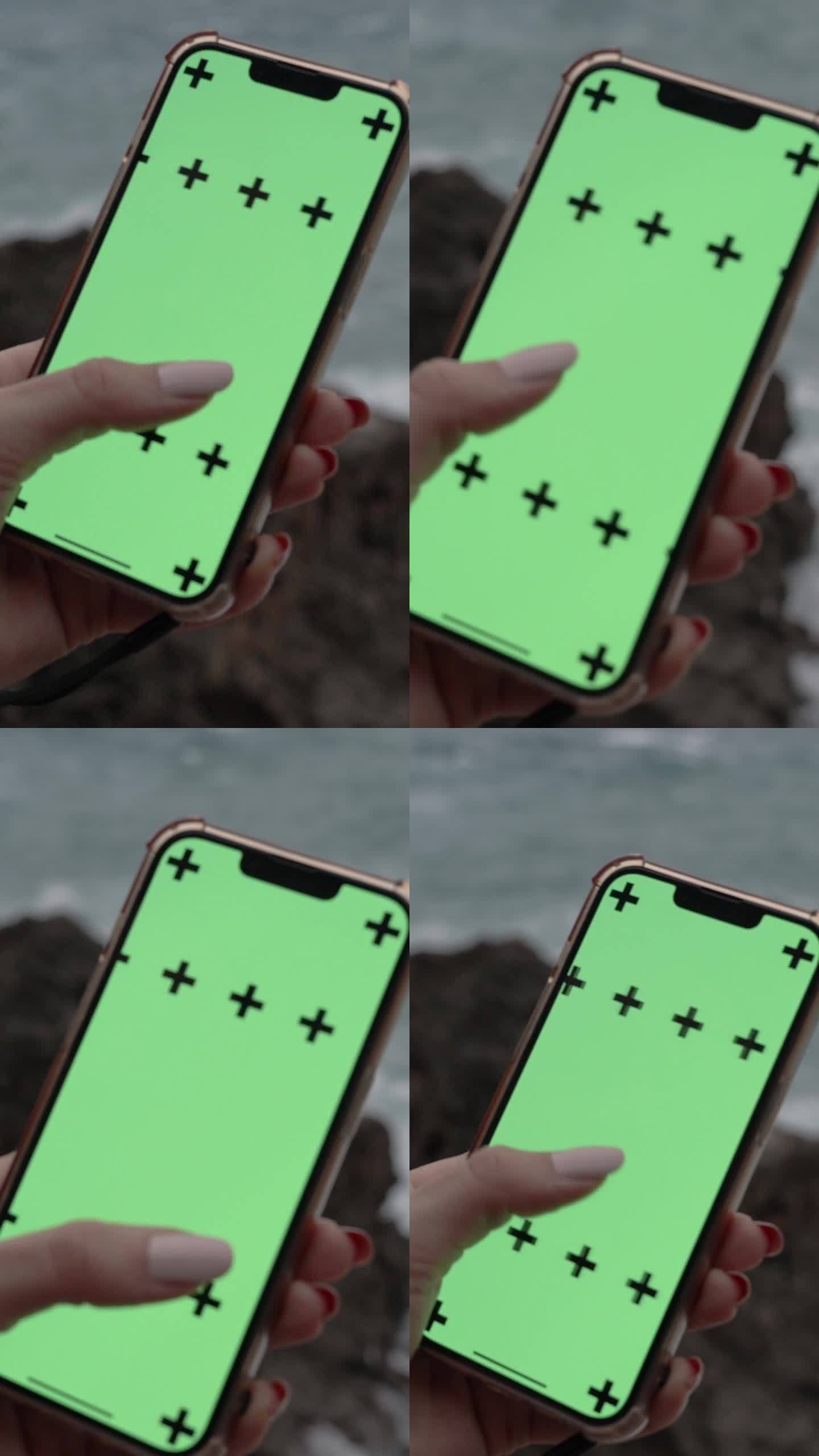 垂直视频。镜头拉近了智能手机，一个女人在看照片，屏幕上是一个空的绿色屏幕。背景是岩石和波涛汹涌的大海