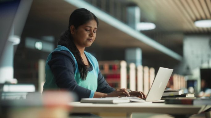 一名印度女性在公共图书馆使用笔记本电脑上的在线办公软件，专心完成一项大学项目的课堂作业。年轻的南亚妇