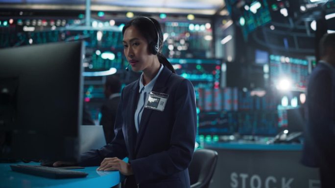 一位在国际证券交易公司工作的年轻亚洲女性的肖像:交易员站在桌子后面，带着电脑，戴着耳机与公司业务伙伴