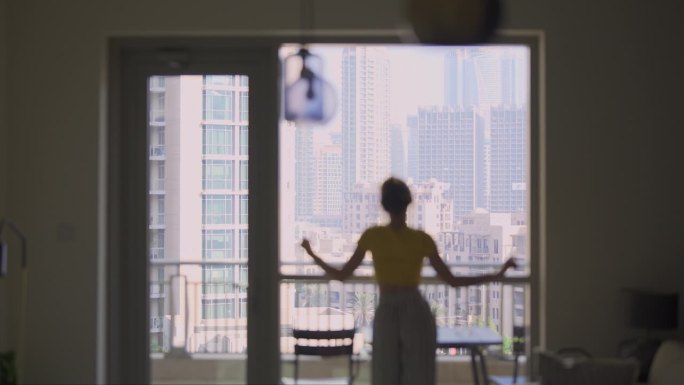一名女子站在窗边，从她的公寓里看着迪拜的城市景观