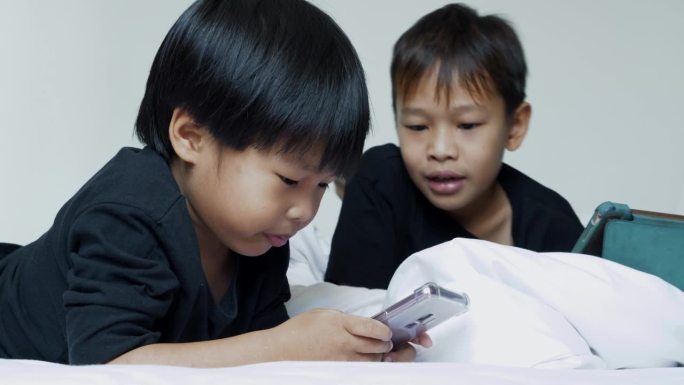 亚洲兄弟男孩躺在床上开心地在家玩手机，家庭纽带概念。