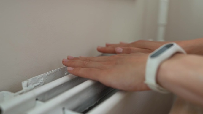 女性手放在暖气片上的特写。女性把手放在家里的加热器上。女人把他的手放在暖气片上暖手。在寒冷的冬季使用