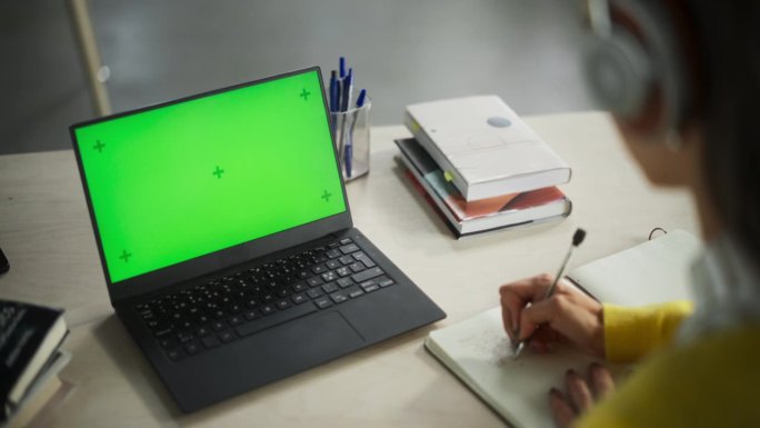 女性在绿色屏幕模拟显示的笔记本电脑上听在线讲座，并就纸质笔记本做笔记。在公共图书馆学习的大学生。匿名