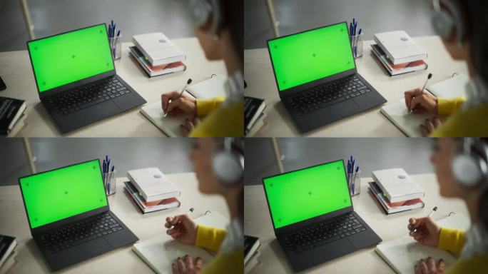 女性在绿色屏幕模拟显示的笔记本电脑上听在线讲座，并就纸质笔记本做笔记。在公共图书馆学习的大学生。匿名