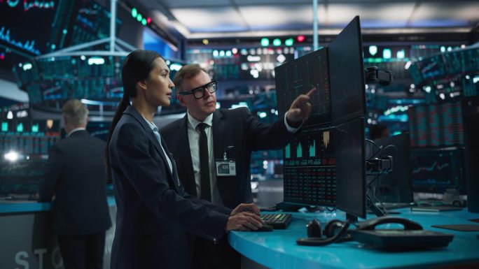 亚洲男女证券交易代理人使用电脑，为国际商业客户讨论投资策略，在被电脑屏幕包围的办公大楼里工作