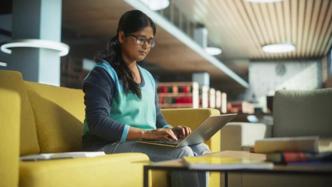 年轻的印度女性专家在一家国际创业公司办公室的休息区工作。项目经理把她的笔记本电脑放在膝盖上，浏览互联