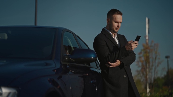 一位穿着大衣的优雅商人站在他的高档轿车旁，用智能手机解决家庭和工作问题。