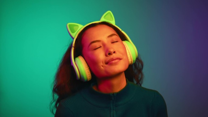 快乐的亚洲女人的肖像，戴着耳机，在霓虹灯下听音乐。一个微笑的女孩听着网络Mp3播放器上的在线歌曲。放