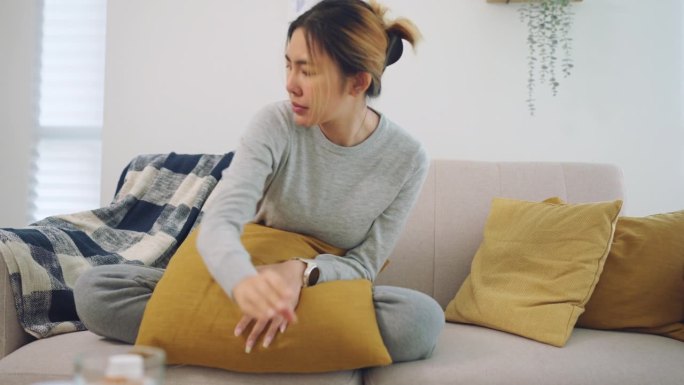 亚洲妇女有流感病毒覆盖格纹在纸巾打喷嚏流鼻涕坐在家里的沙发上恶心，季节性病毒