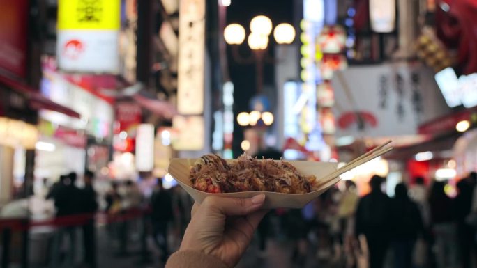 在大阪市中心的街道上，一名女游客正拿着新鲜制作的日本传统街头小吃章鱼烧(章鱼球)。镜头从她的个人角度