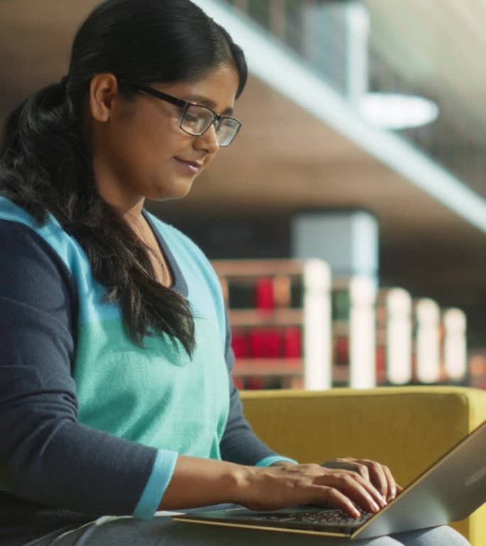 垂直屏幕:印度女性在公共图书馆使用笔记本电脑完成大学项目的课堂作业。年轻的南亚妇女在做练习的同时在线