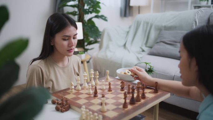 年轻的亚洲女子在家里和朋友下棋时吃着零食