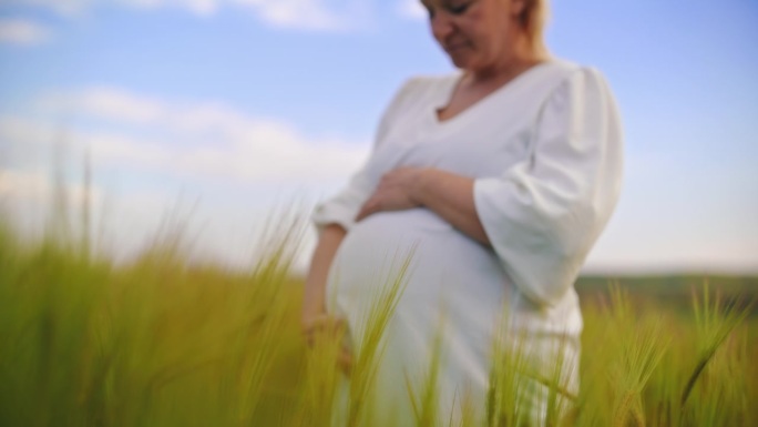 近距离拍摄身穿白色连衣裙的孕妇在田野里抱着肚子