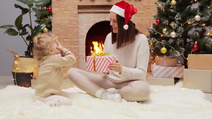 圣诞祝福的季节。快乐的妈妈和可爱的女儿在家里的装饰圣诞树和壁炉旁玩耍，美丽的妈妈给婴儿送礼物盒。