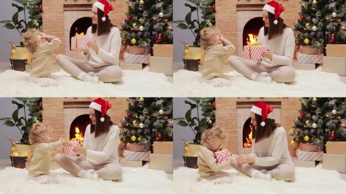 圣诞祝福的季节。快乐的妈妈和可爱的女儿在家里的装饰圣诞树和壁炉旁玩耍，美丽的妈妈给婴儿送礼物盒。