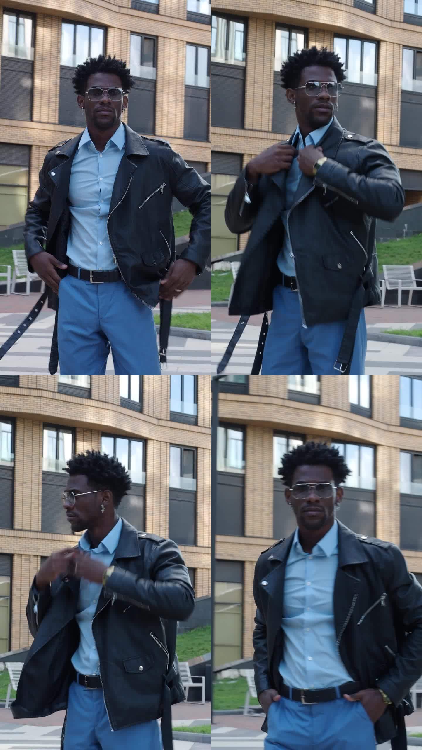 一个黑人年轻人，穿着时髦的皮夹克，蓝色衬衫，裤子，戴着太阳镜，站在城市街道上，看着镜头。帅气男时尚模