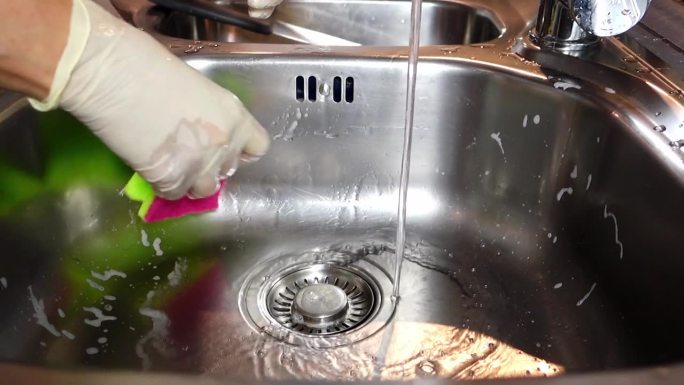 一位妇女小心地冲洗洗碗海绵和洗涤液。紧紧戴着手套的手把海绵从泡沫中拧出来。