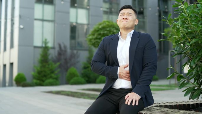 穿着正装的亚洲商人坐在办公大楼附近的长椅上感到胃痛。