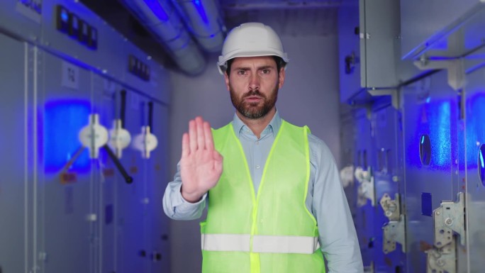 戴白色安全帽的男性电气工程技术工人的肖像，变电所工人在蓝光、高压和故障电气设备的背景下示意停车。