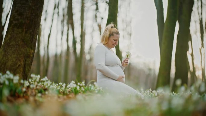 宁静的孕妇坐在树林里白色的雪花莲花中间