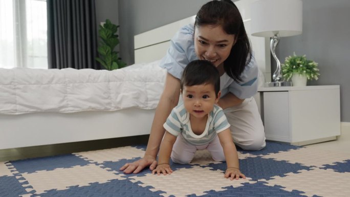 快乐的母亲看着她的婴儿在卧室的游戏垫或拼图地板上爬行