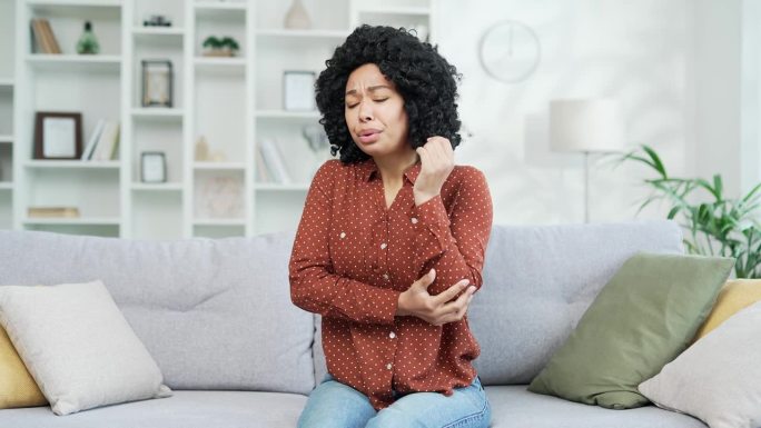 年轻的非裔美国女性穿着衬衫坐在家里客厅的沙发上，肘部疼痛。