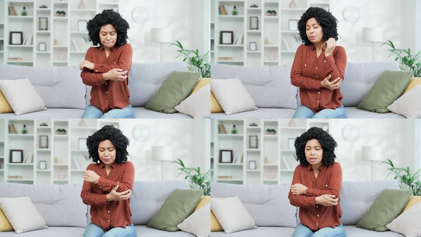 年轻的非裔美国女性穿着衬衫坐在家里客厅的沙发上，肘部疼痛。