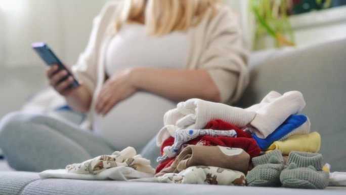 孕妇叠婴儿衣服，在沙发上玩智能手机