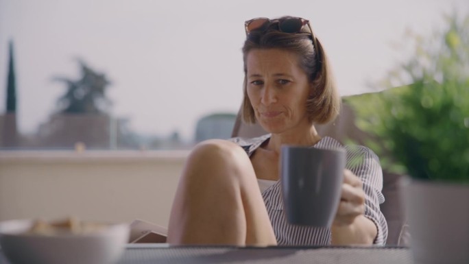 手持拍摄的中年成年妇女一边喝咖啡，一边在家里的露台上看书
